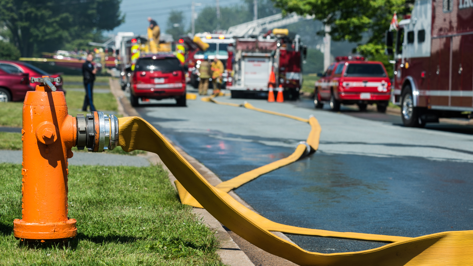 Orange fire hydrant alongside a street, with fire hose hooked up to it and fire trucks on the street in the background.